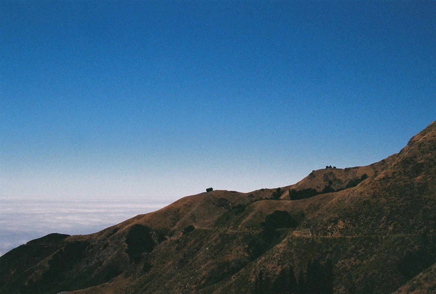 A scenic landscape photograph of rolling brown and green hills under a clear blue sky, with a layer of low white clouds on the horizon.