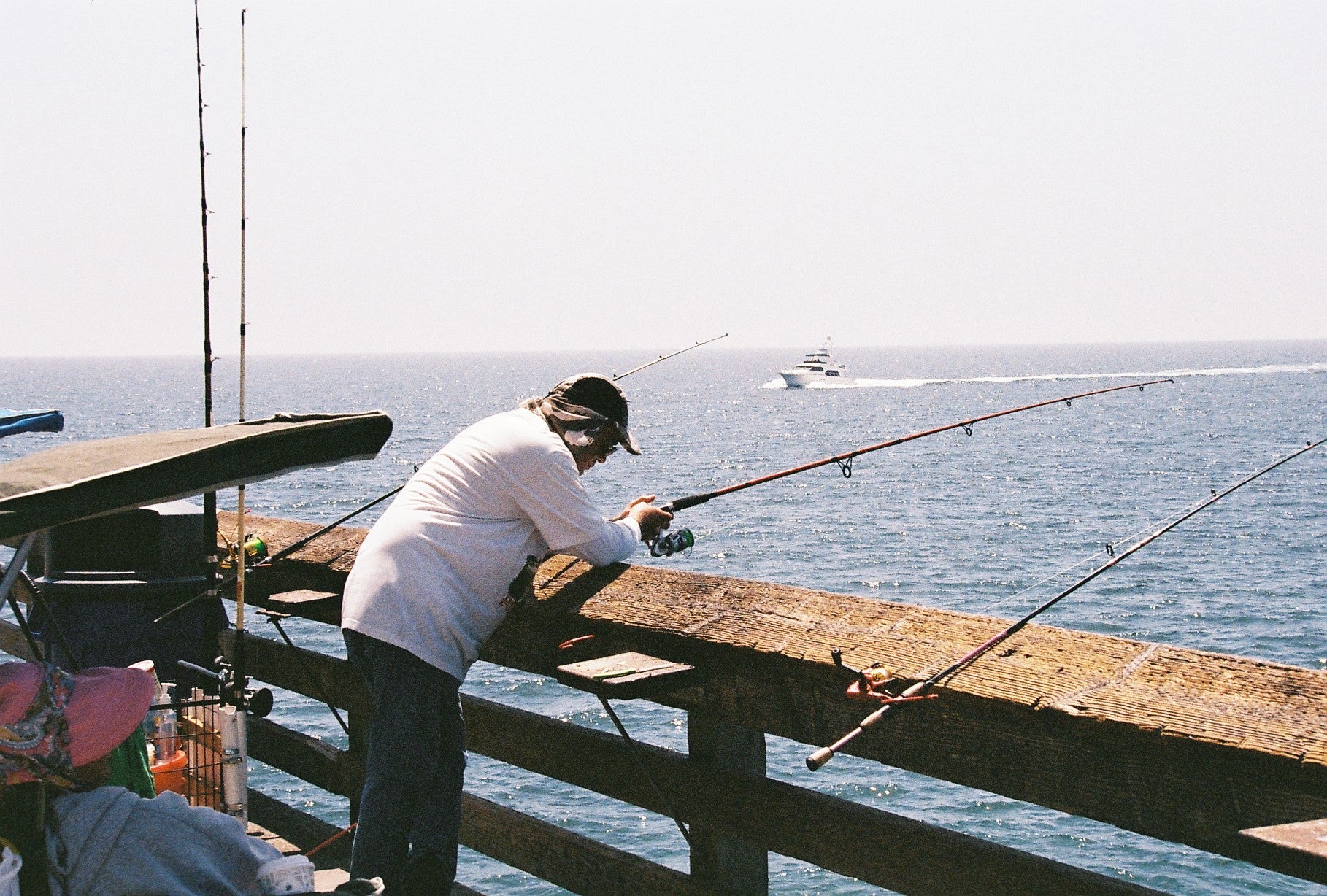 A person in a white long-sleeve shirt and dark pants fishes from a wooden dock over the ocean, with fishing rods and a boat visible in the water under a bright sky.