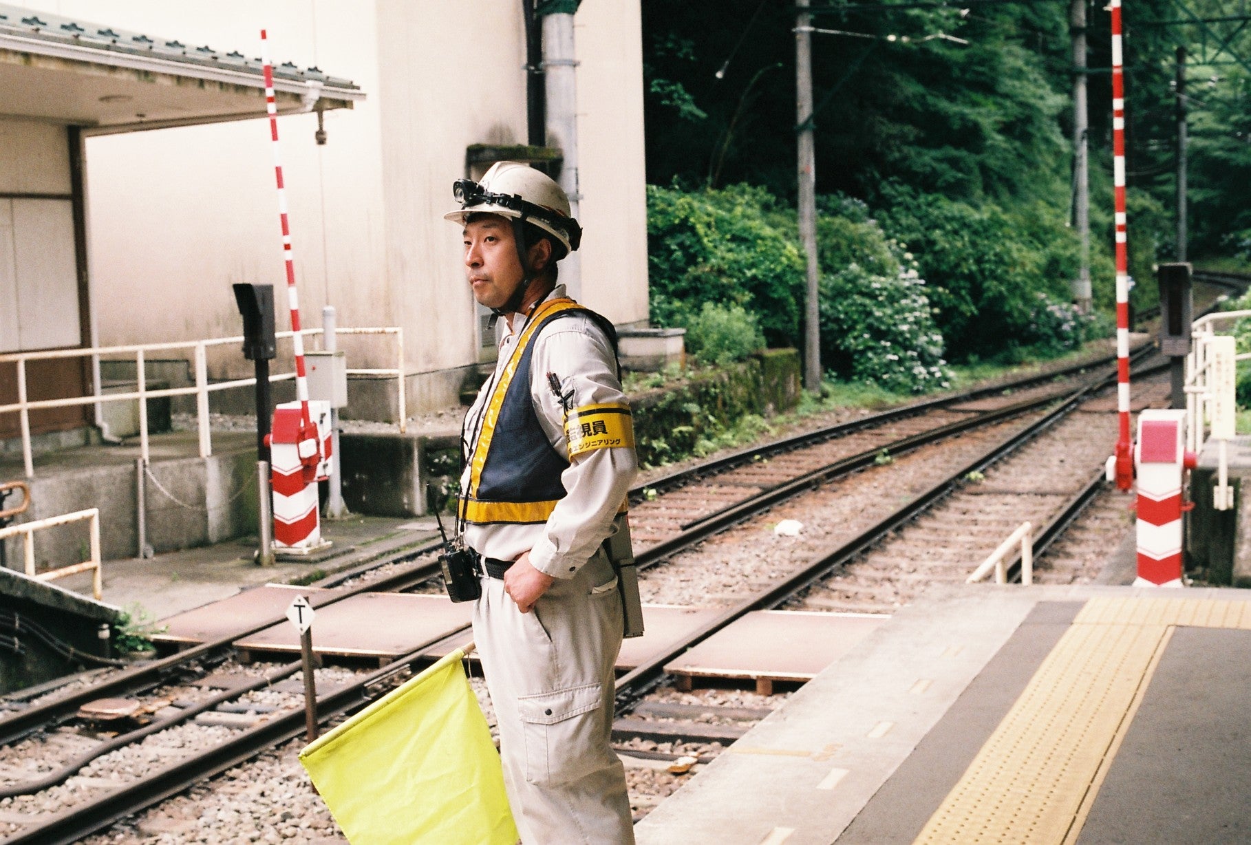 A railway signalman in a light gray uniform and white hard hat stands on a platform next to train tracks, holding a yellow flag. The background features train tracks, green trees, and signal barriers.